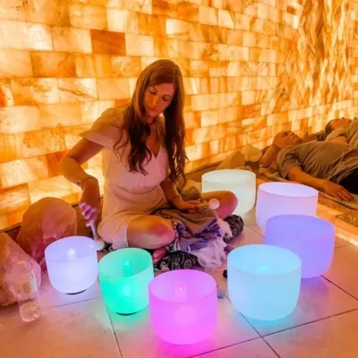 People laying in the back by a woman sitting in front of a LED backlit salt panel with sound therapy bowls lit with colored lights.