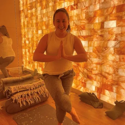 Woman in the tree pose on a toga mat in front of a mirror and an LED backlit salt panel wall at Sweet Be Wellness and Skin Clinic - Greeley, Colorado.