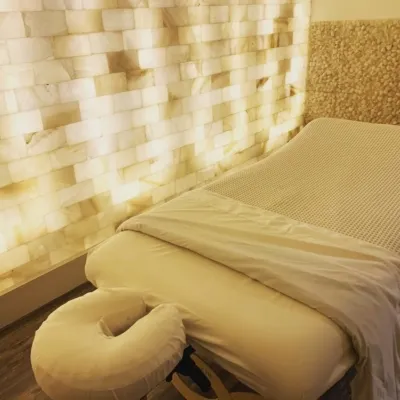 White massage table on a brown vinyl flooring in front of an LED backlit Himalayan salt panel wall.