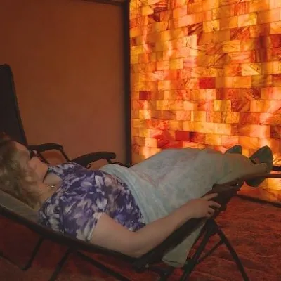Women reclined relaxing in a Salt Spa on a salt-covered floor facing an orange backlit Himalayan salt stone paneled wall.