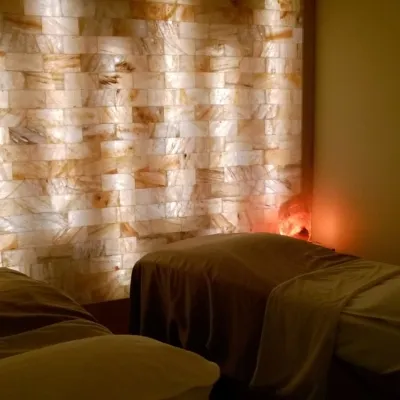Two massage tables with pillows and blankets in a small room with a LED backlit salt brick wall. There is a round mirror hanging on the wall to the right, and a lit Himalayan salt lamp propped up on a table in the corner of the room.