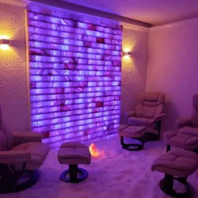 Three tan chairs with ottomans in a salt-covered room with a purple LED backlit salt brick wall with a salt rock lamp in the middle of the room