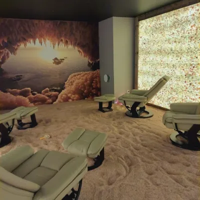 Multiple tan chairs and ottomans in a room with a salt-covered floor, an LED backlit salt wall on the right and a mural of a salt cave on the left.
