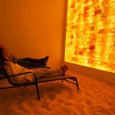 A woman and child in two lounge chairs in a dimly lit salt room with salt-covered floors facing a LED backlit salt brick wall