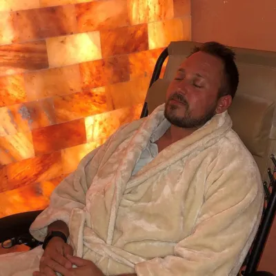 Sweet Be Wellness and Skin Clinic. Man in white robe laying back in reclining chair with eyes closed in salt room