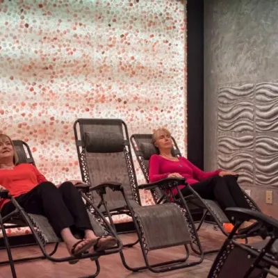 The Acupuncture Center and salt spa. Two women sit in lounge chairs with a large salt, stone wall behind them. One woman is in a salmon colored shirt while the other is in a magenta shirt.