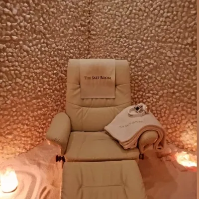 The Salt Room. Recliner in corner of salt room. Headphones on top of towels lay on the chair and a cloth is draped over the back of the chair that reads "The Salt Room."