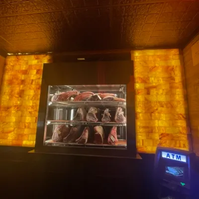 A glass fridge of meat The Market Tavern in Flanders, New Jersey in front of an orange backlit salt stone wall with an ATM on the side.