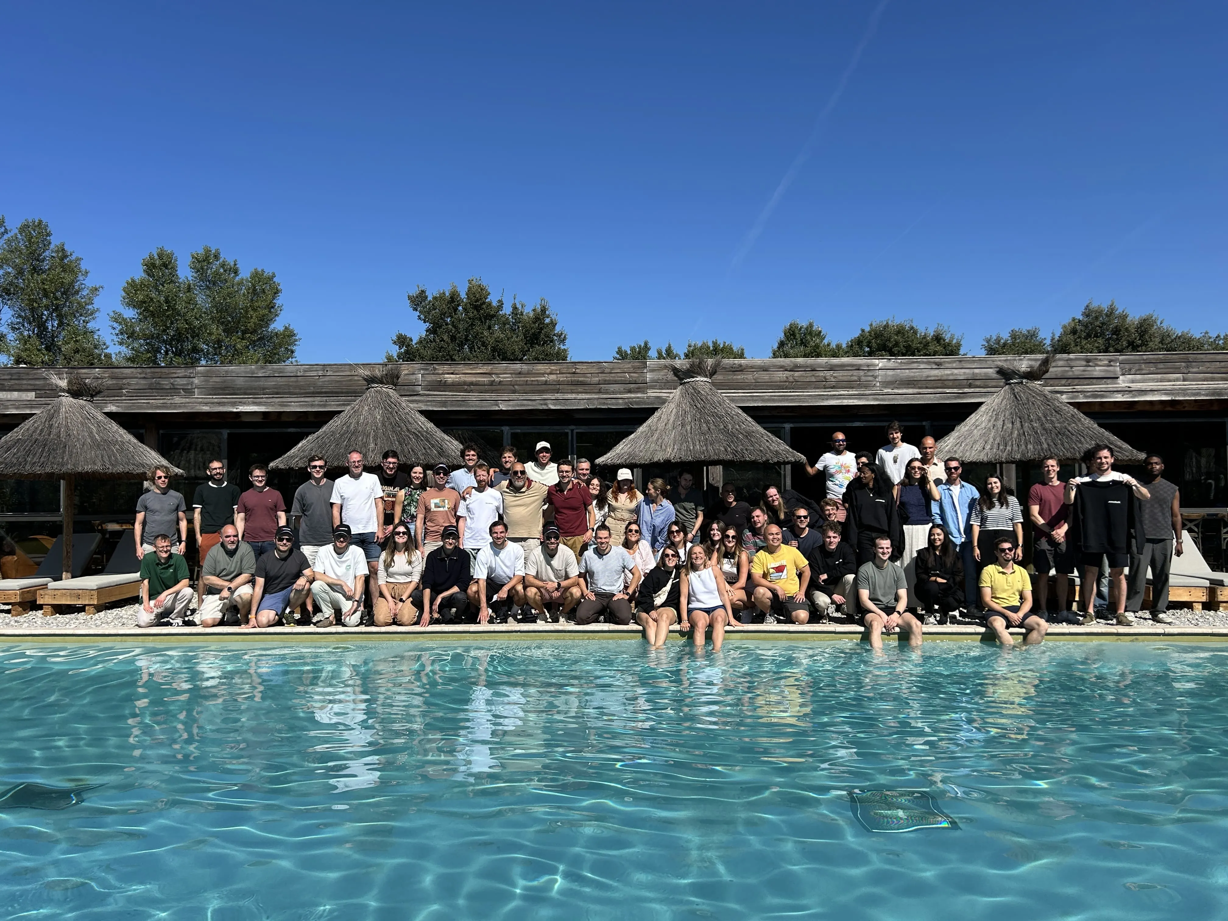Large group of people posing together by a poolside with thatched umbrellas and a wooden building in the background under a clear blue sky.