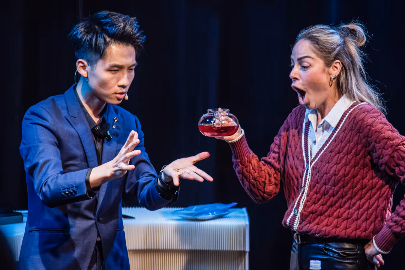 Man in blue blazer performing a magic trick with a spider as a woman in a red sweater watches shocked holding a glass teapot with red liquid.