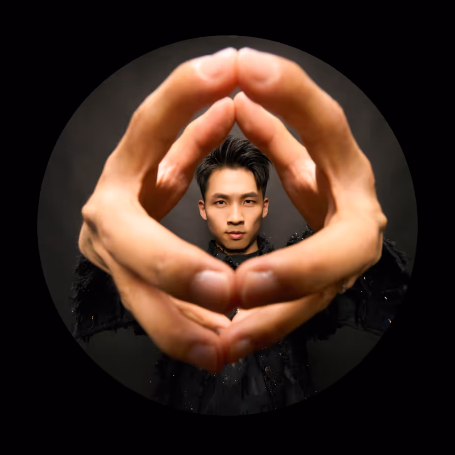 Man with dark hair making a circular frame with his hands in front of his face, against a black background.
