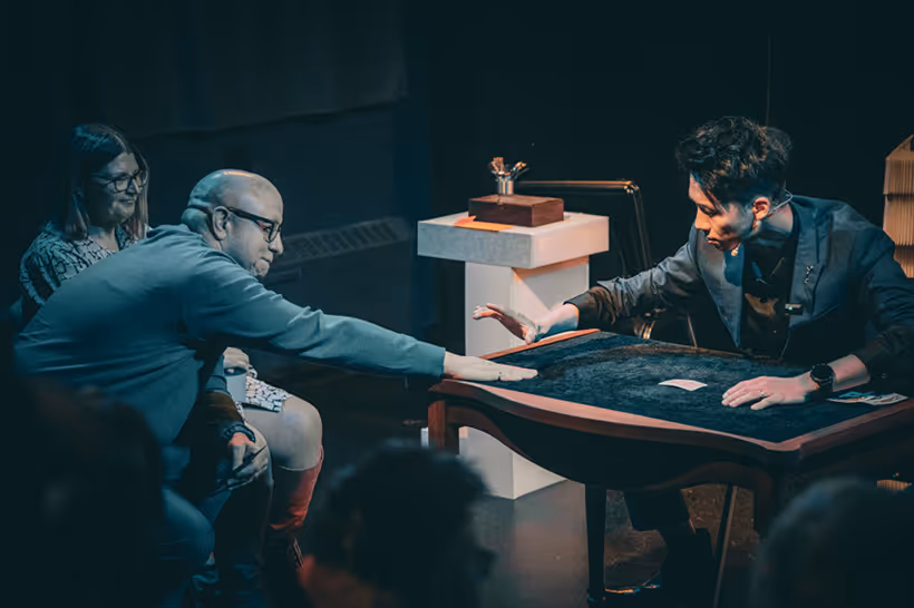 Man in glasses reaching out to touch cards on a table while a magician gestures during a close-up magic performance.
