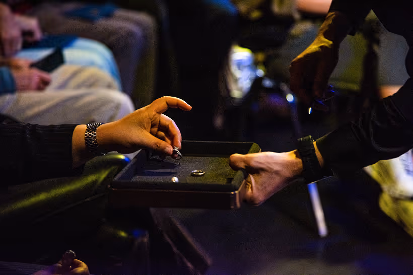 Two hands exchanging a black tray with rings in a dimly lit setting with seated people in the background.