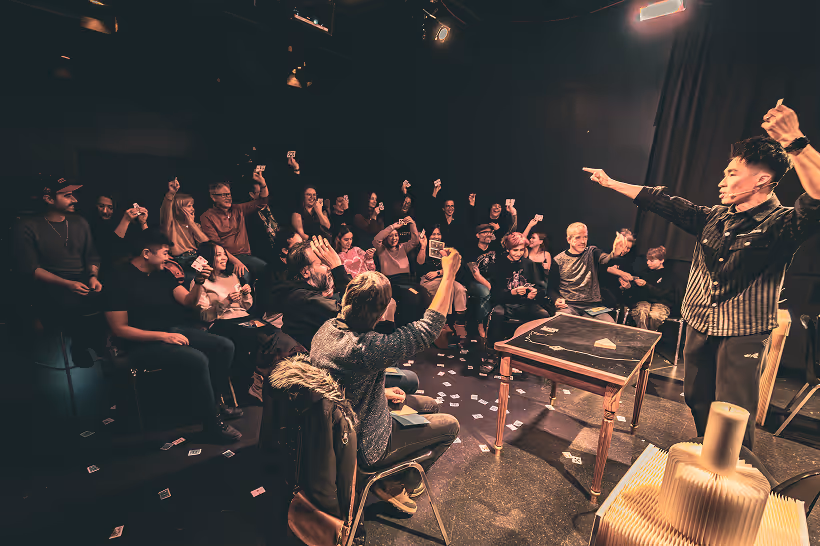 Magician performing card tricks in front of a seated audience who are enthusiastically holding playing cards.