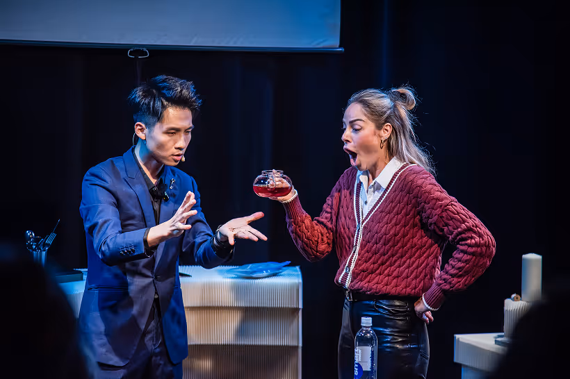 Man in blue suit performing a magic trick with a floating glass teapot while a woman in a red sweater reacts with surprise.