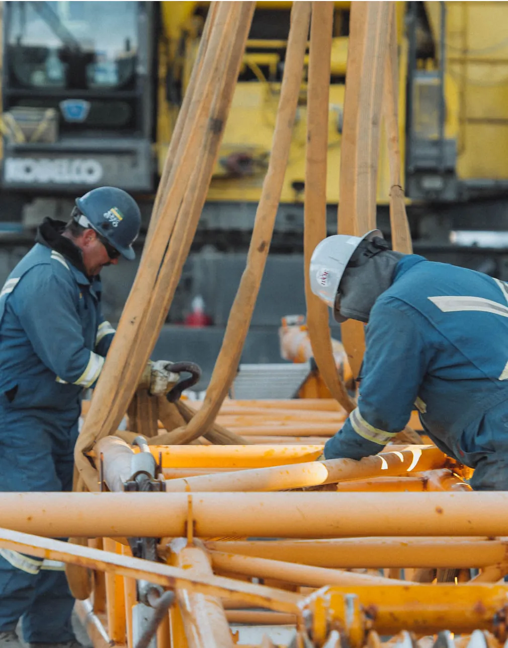 two men in blue work on a construction site with a yellow crane