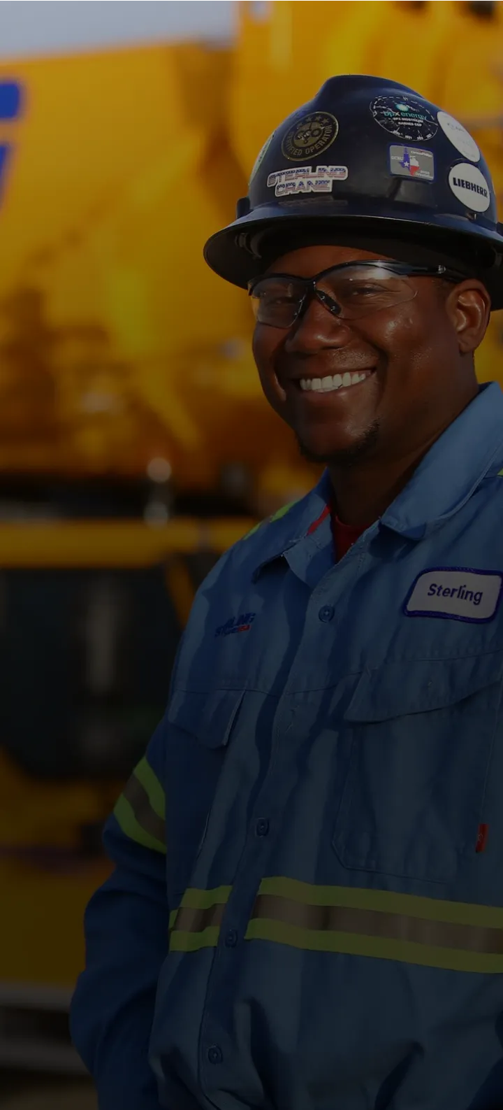 smiling man in blue workwear and hard hat standing in front of a yellow truck