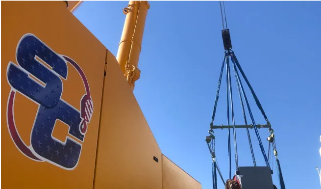 crane lifting a large yellow construction vehicle with a blue sky in the background