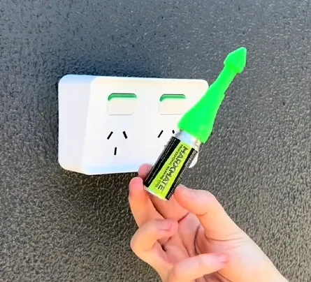 A hand holding a green Marxmate marking tool in front of a white double power outlet mounted on a textured grey wall.