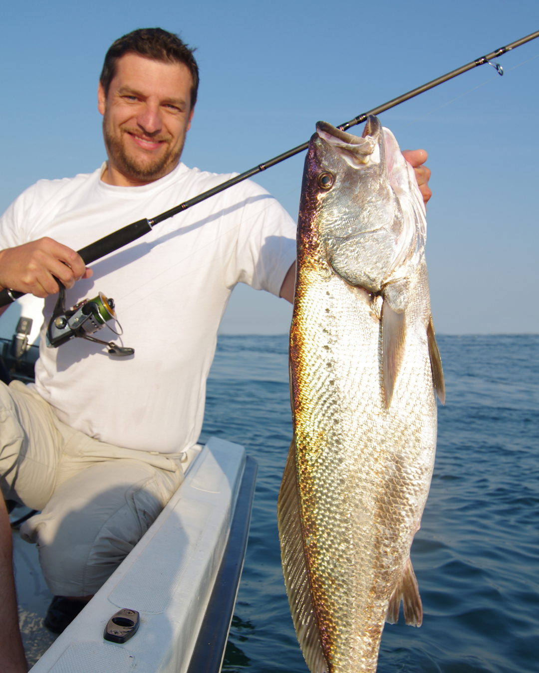 Smiling angler on a boat holding a large fish beside a spinning rod