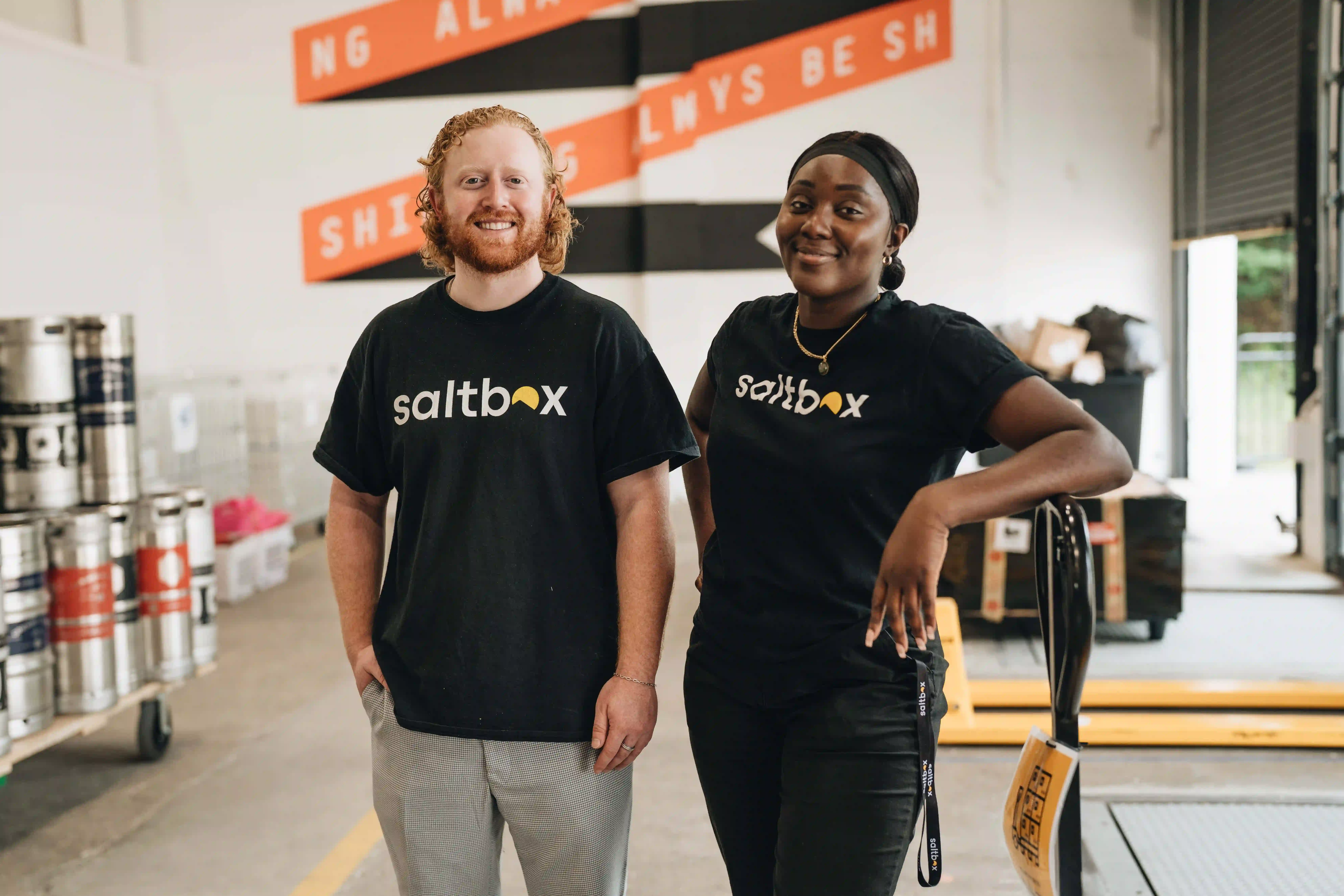 Two Saltbox employees smiling and standing in a warehouse with kegs and equipment in the background.