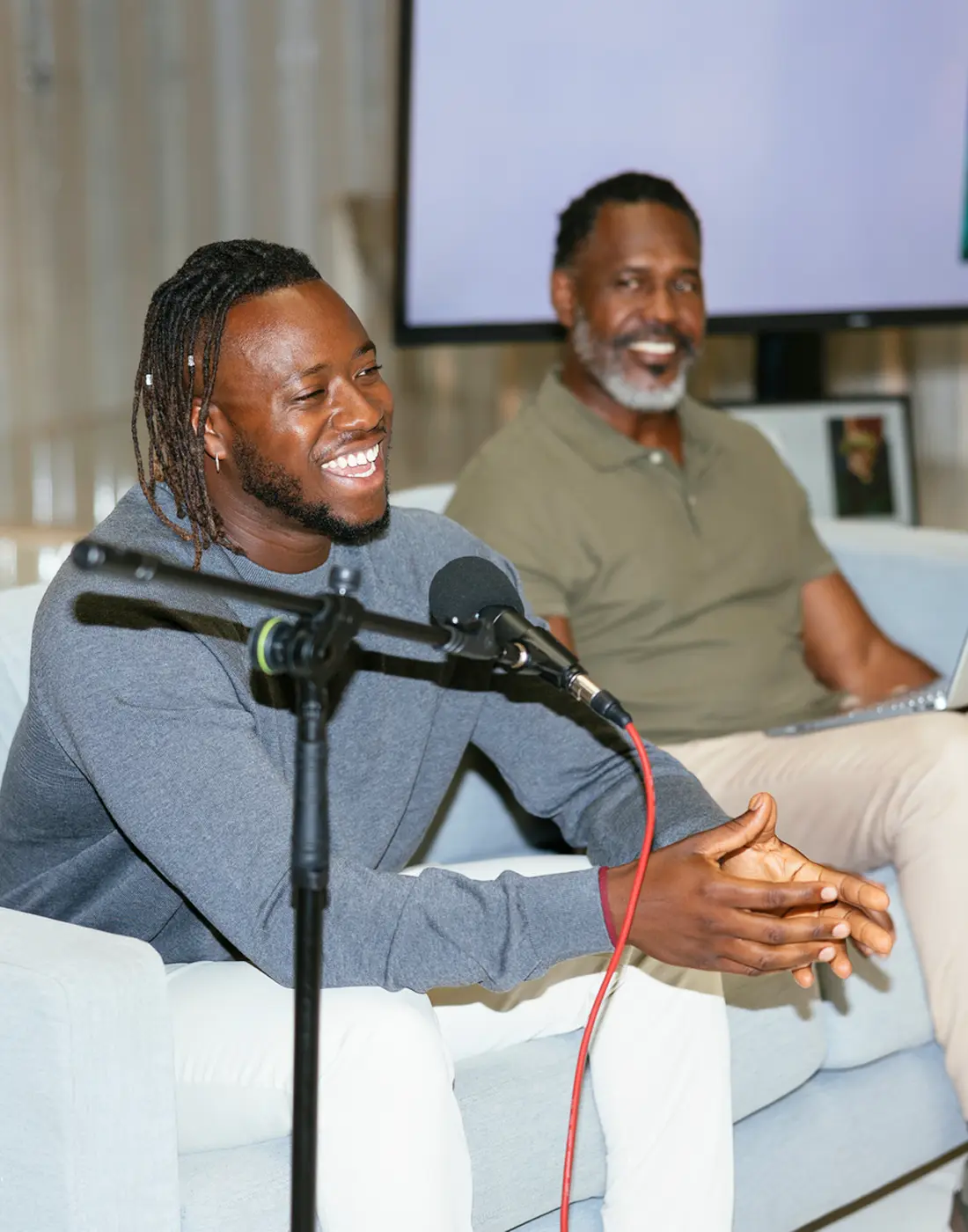 Two men sitting on a couch, one speaking into a microphone and laughing, the other smiling with a laptop on his lap.