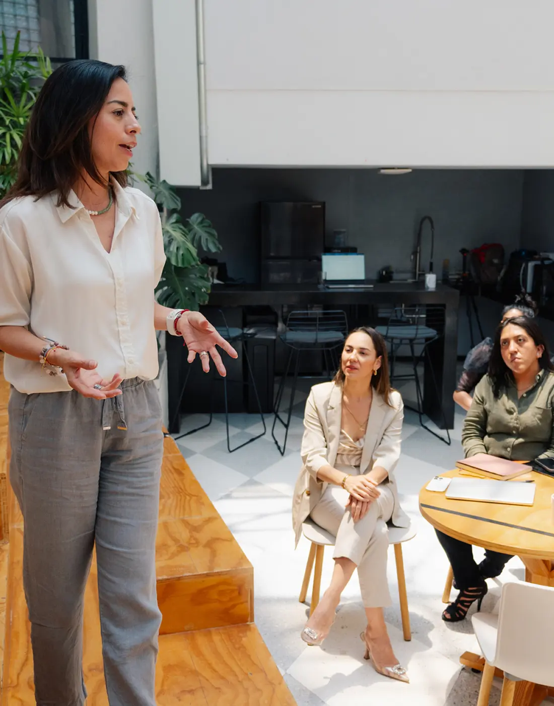 Woman in casual attire speaking to two seated women attentively listening in a modern office space.