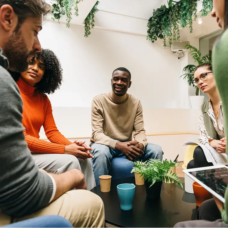A diverse group of five people sitting around a table having a discussion in a bright room with plants.