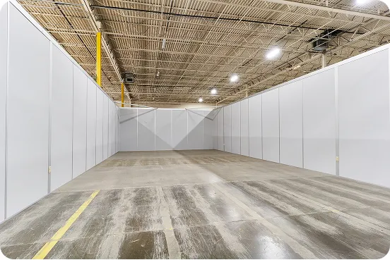 Empty large warehouse space with concrete floor and tall white partition walls under bright ceiling lights.