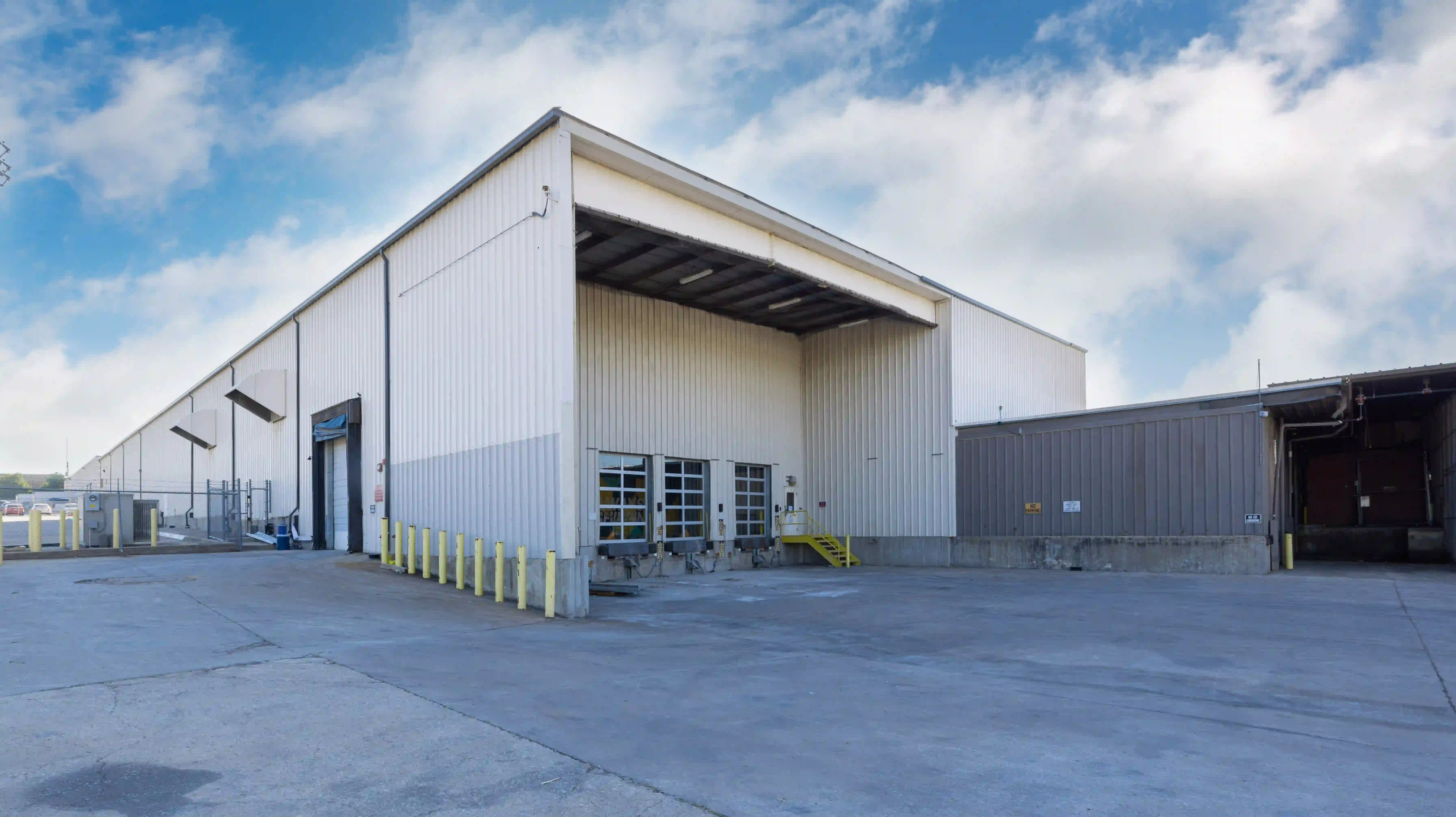 Large industrial warehouse with multiple loading docks and yellow safety bollards under a partly cloudy sky.