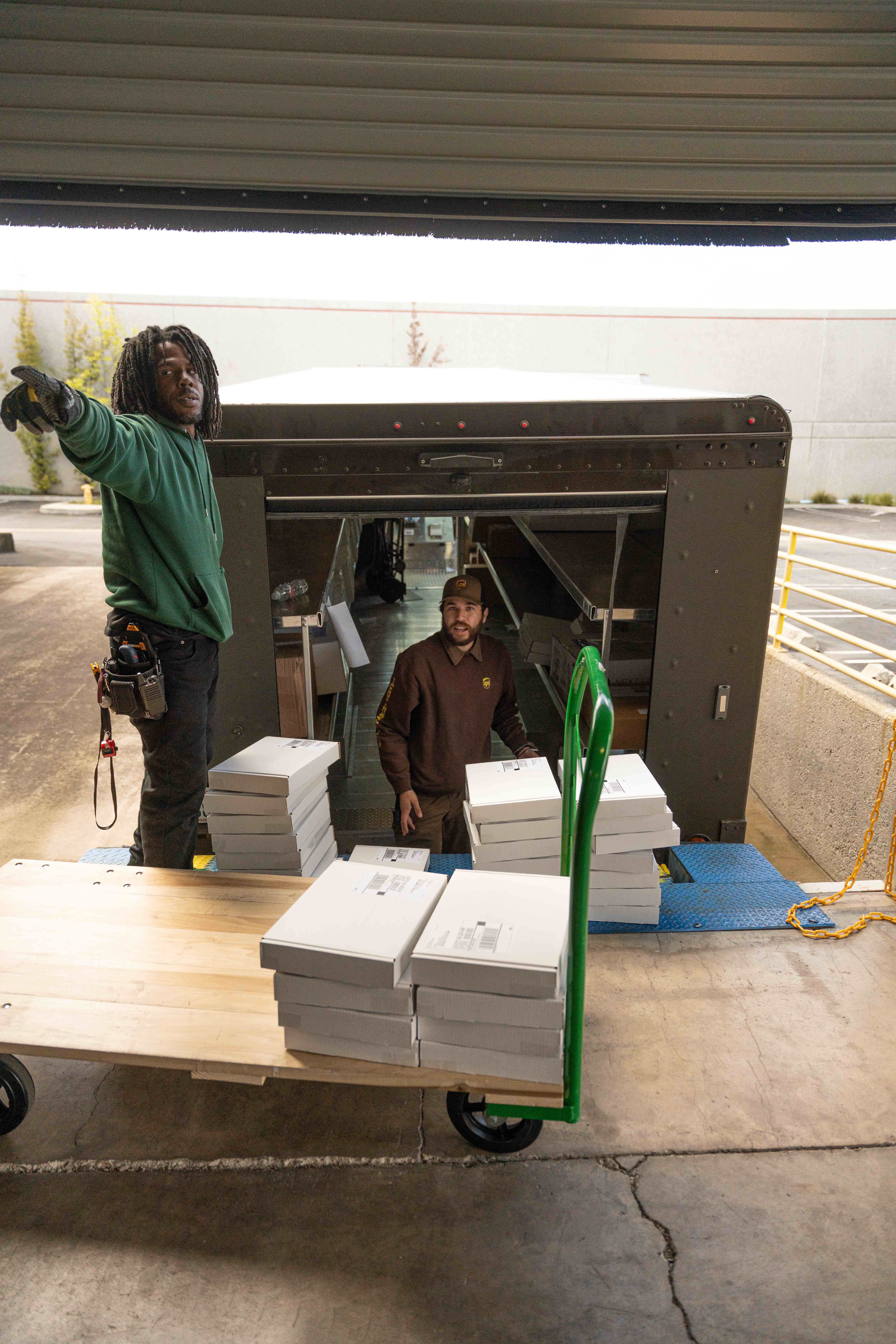 Two delivery workers handling stacks of white boxes near the open back of a delivery truck with a loaded cart.