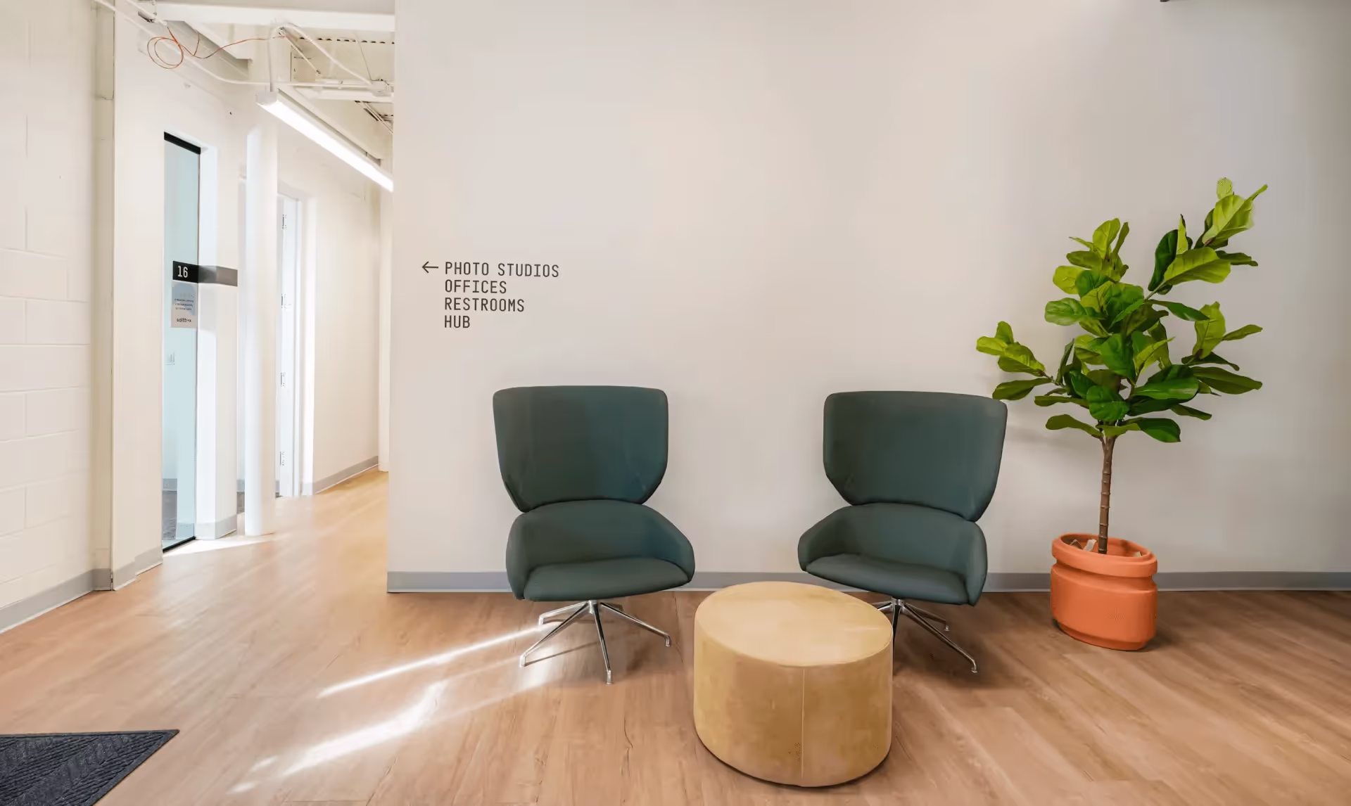 Modern seating area at Saltbox with two green chairs, a small round table, and a potted plant near the hallway to offices, photo studios, and restrooms