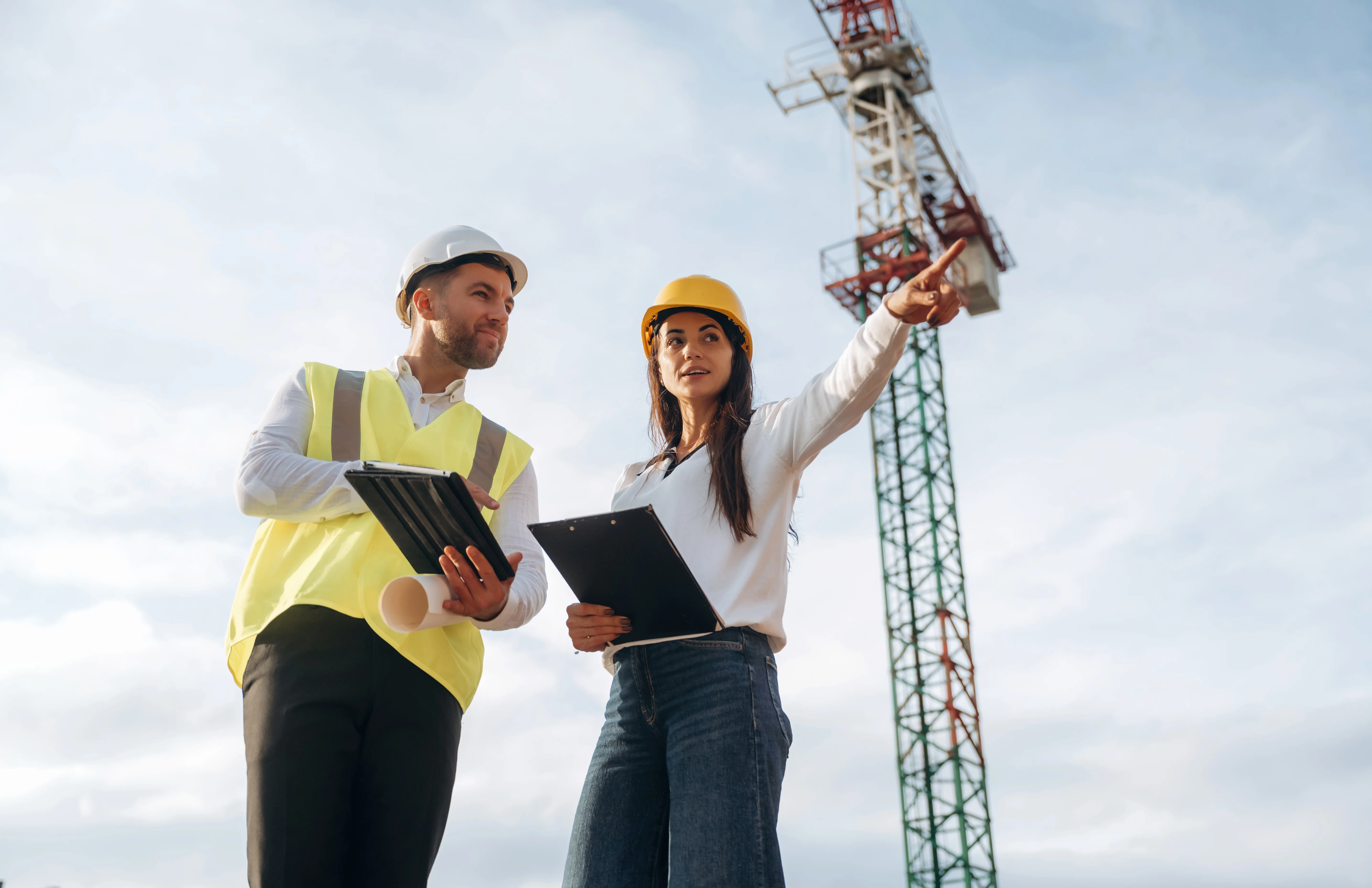 Two utility professionals in high-visibility safety vests and hard hats standing in a field of tall grass, with one pointing toward the horizon while the other holds a digital tablet.