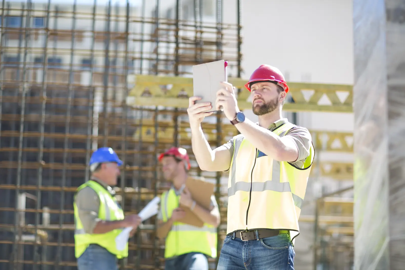  A construction manager in a hard hat and safety vest standing on a high-elevation scaffolding, using a tablet to take a photo or scan of a data center construction site.