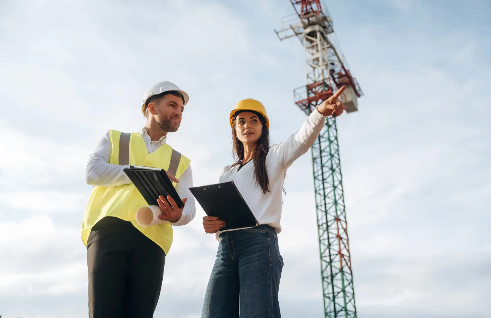 Two utility professionals in high-visibility safety vests and hard hats standing in a field of tall grass, with one pointing toward the horizon while the other holds a digital tablet.