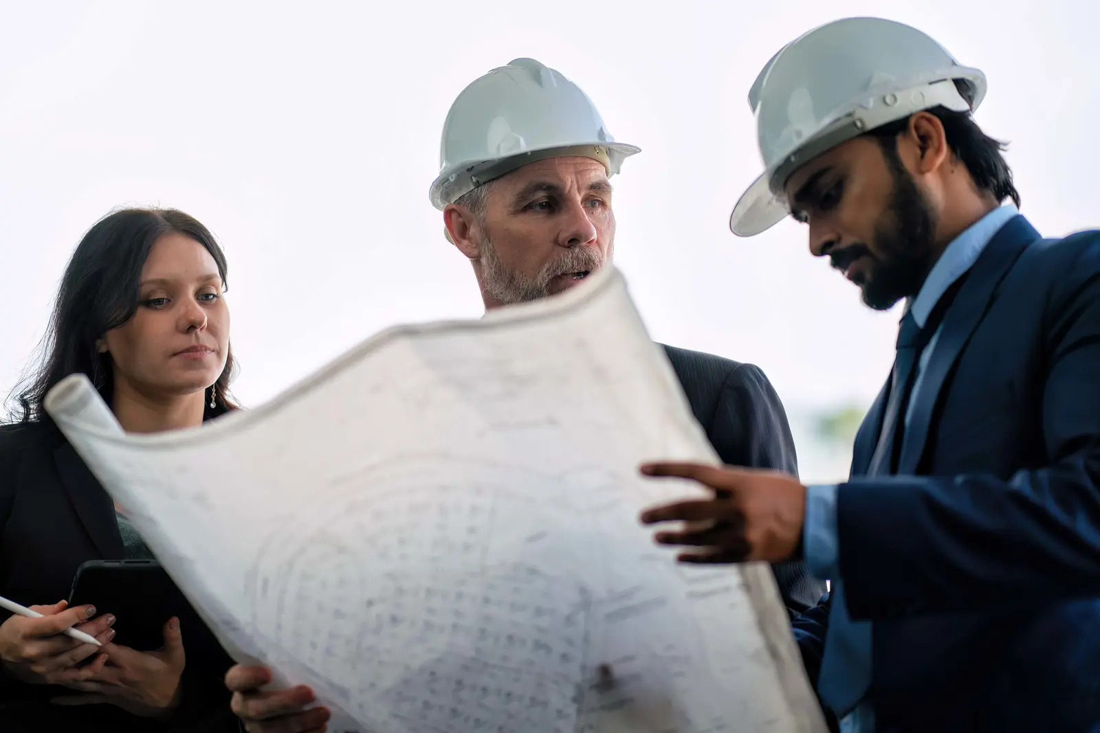 Three construction professionals—a woman in a black blazer and two men in white hard hats and ties—reviewing a large set of blueprints together outdoors.