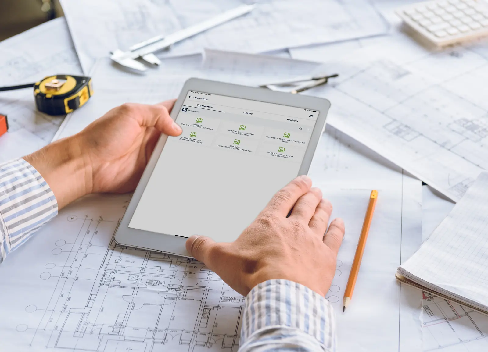 An overhead view of a construction professional’s hands using a stylus on a digital tablet to review architectural blueprints, with a notebook, pen, and white hard hat nearby.