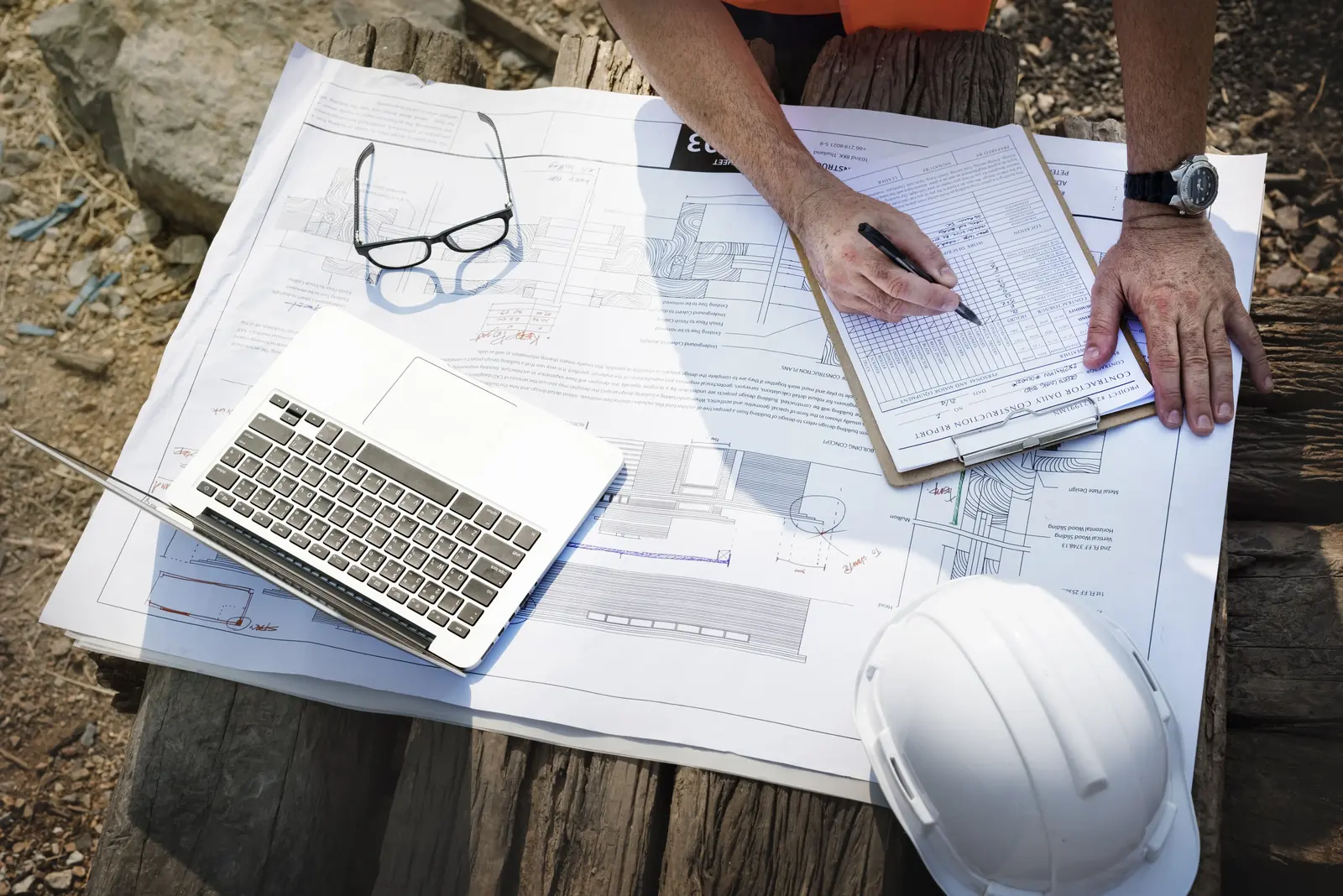 An overhead view of a construction professional’s hands working on a large set of blueprints spread across a table, featuring a laptop, a notebook, and a white hard hat.