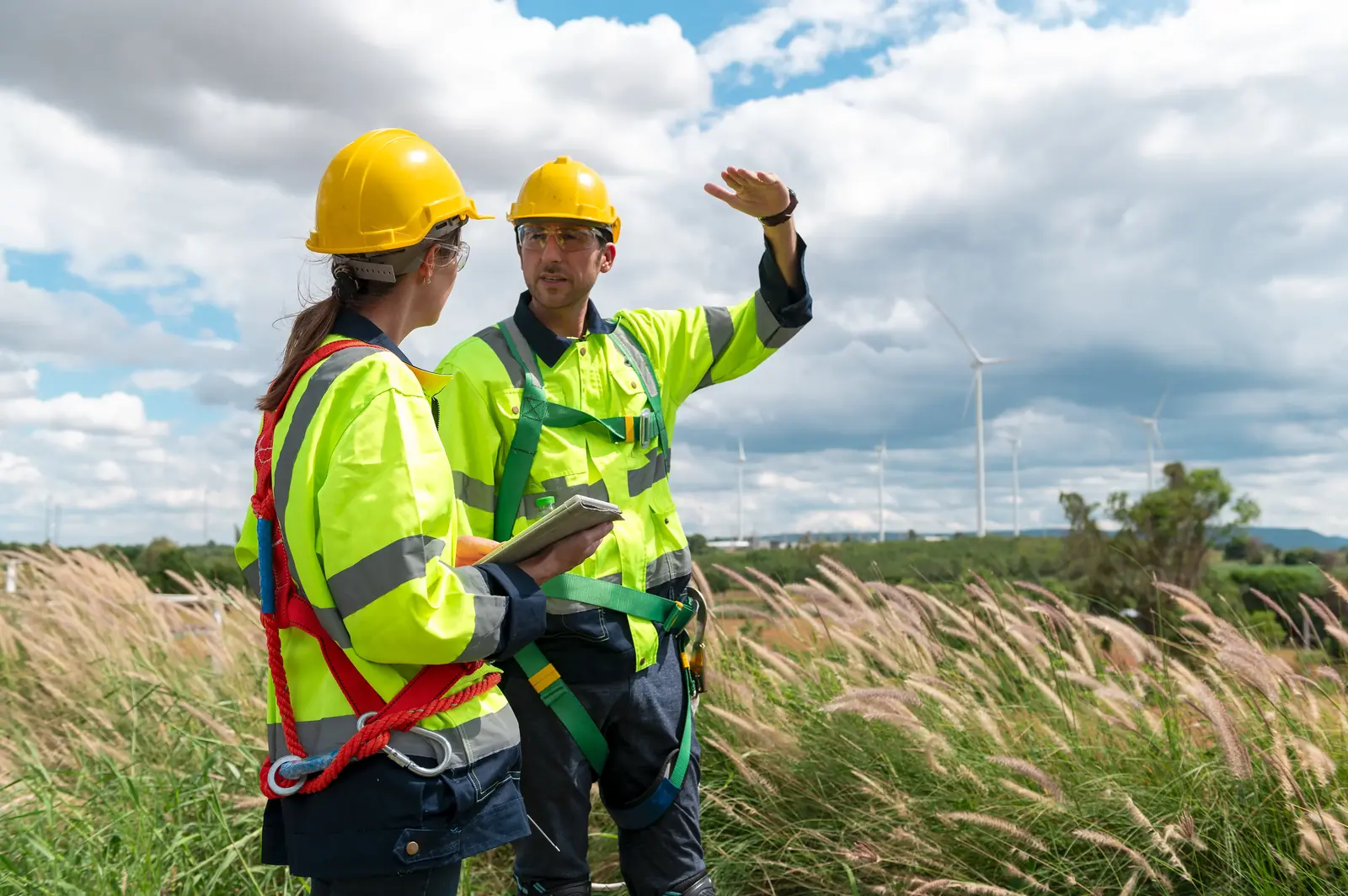 Two utility professionals in high-visibility safety vests and hard hats standing in a field of tall grass, with one pointing toward the horizon while the other holds a digital tablet.