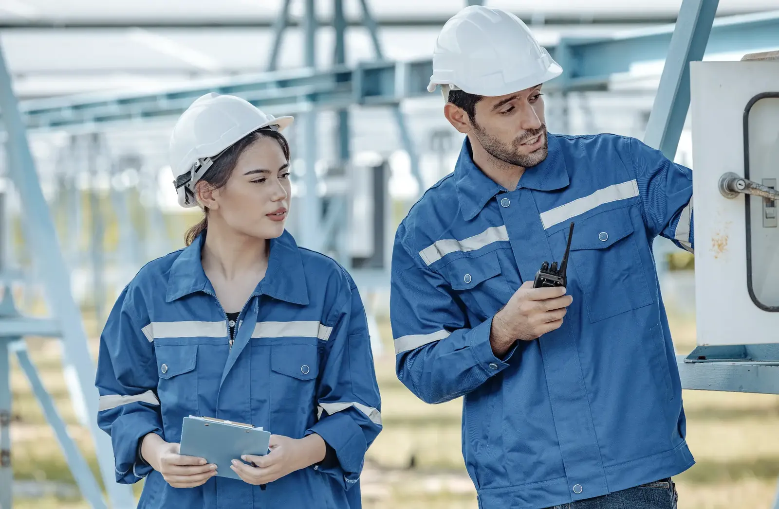 Two utility professionals in high-visibility safety vests and hard hats standing in a field of tall grass, with one pointing toward the horizon while the other holds a digital tablet.
