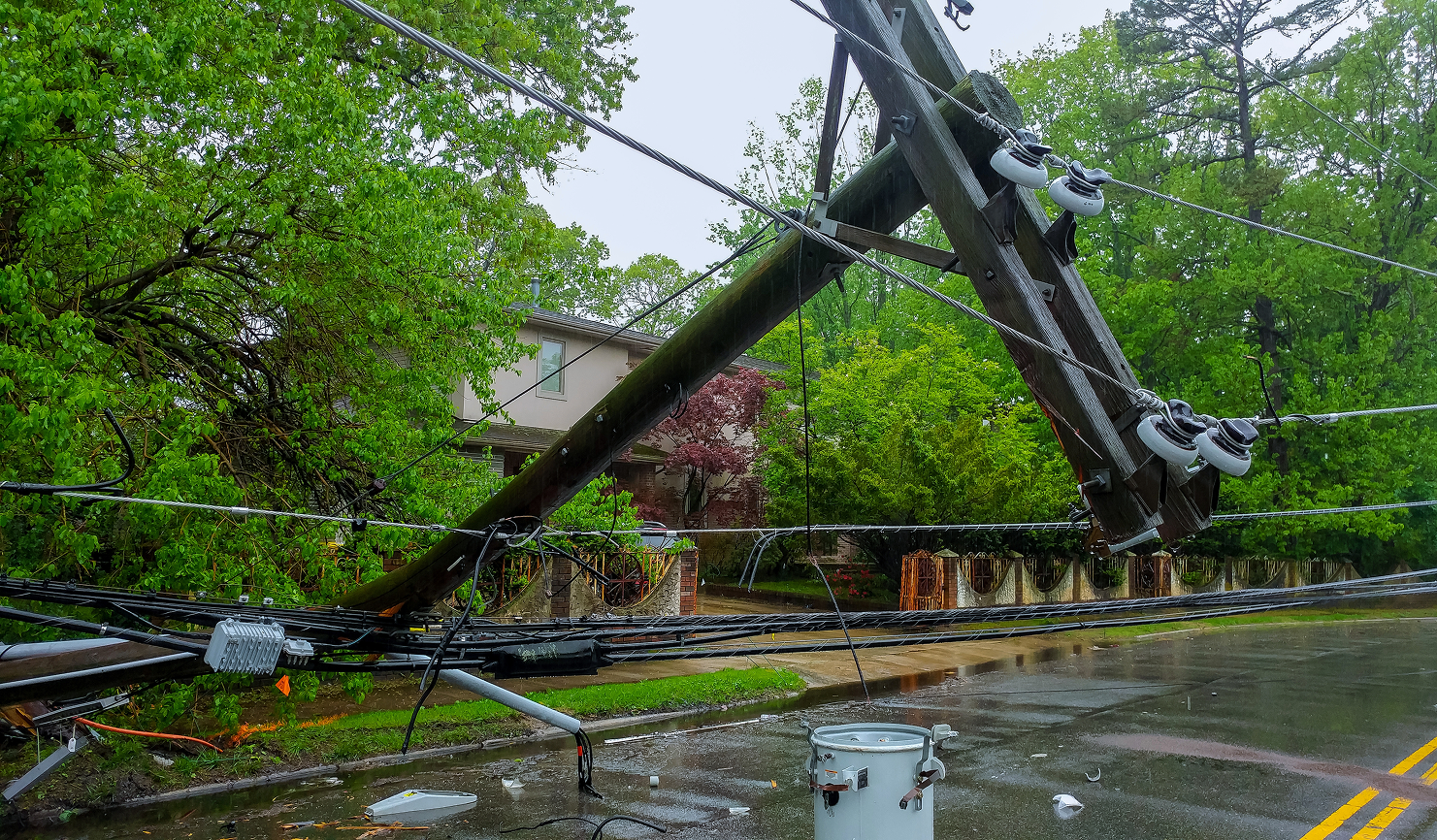 A wooden utility pole has broken and fallen across a suburban street, bringing down power lines and blocking traffic. Lush green trees line the street in this daytime shot. The roadway is damp and there is standing water along the curb line, suggesting it has recently rained. In the background a house can be seen surrounded by trees. This scene is perfect for illustrating weather damage, utility infrastructure issues, and the aftermath of a storm. 