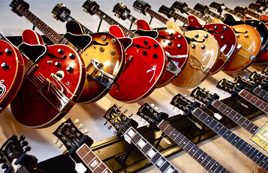 Collection de guitares électriques suspendues en rangée avec différentes finitions rouges, jaunes et naturelles, exposées dans un magasin ou une salle d'exposition de musique.