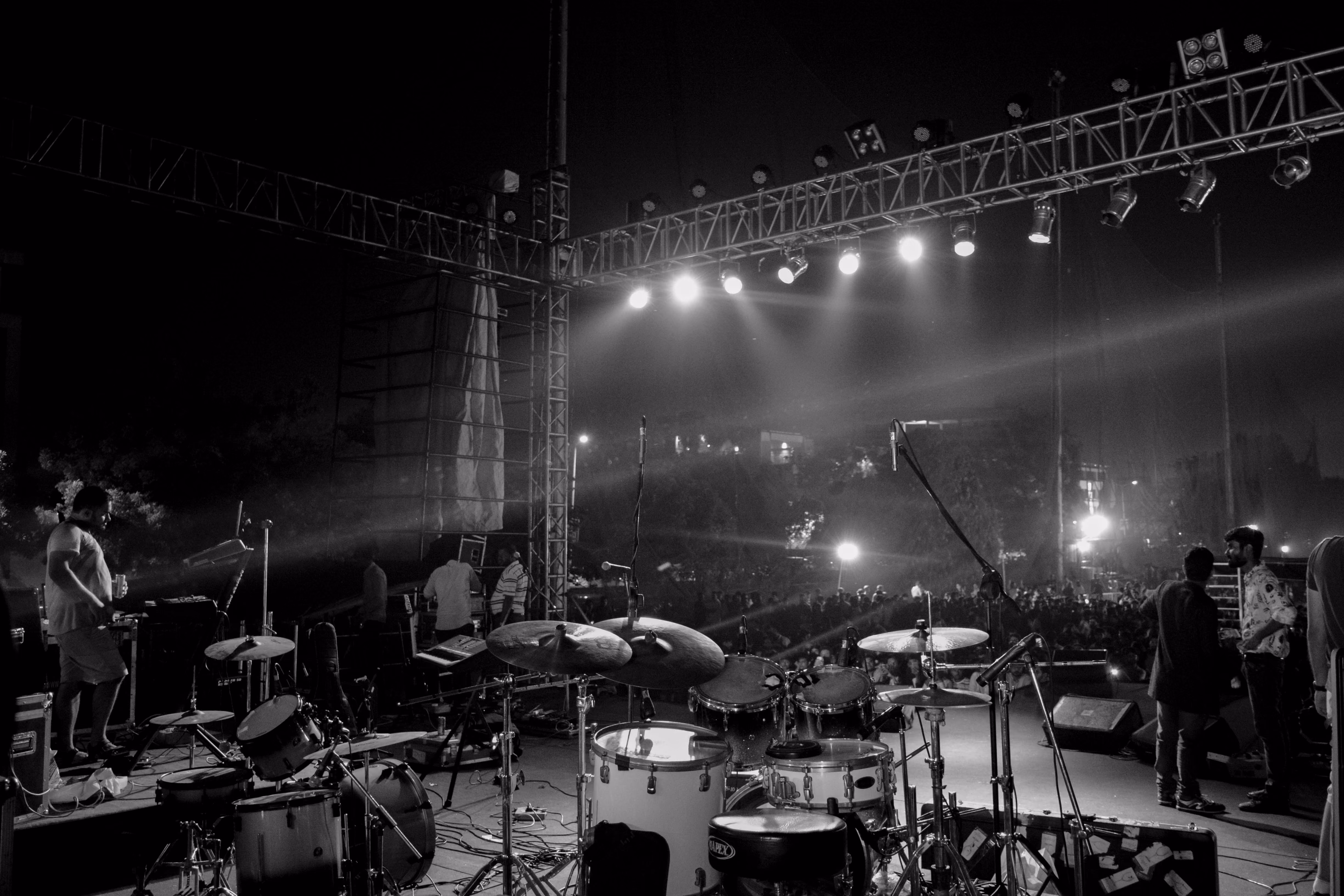 Scène de concert en noir et blanc vue depuis derrière la batterie avec des musiciens et une foule nombreuse sous des projecteurs lumineux.