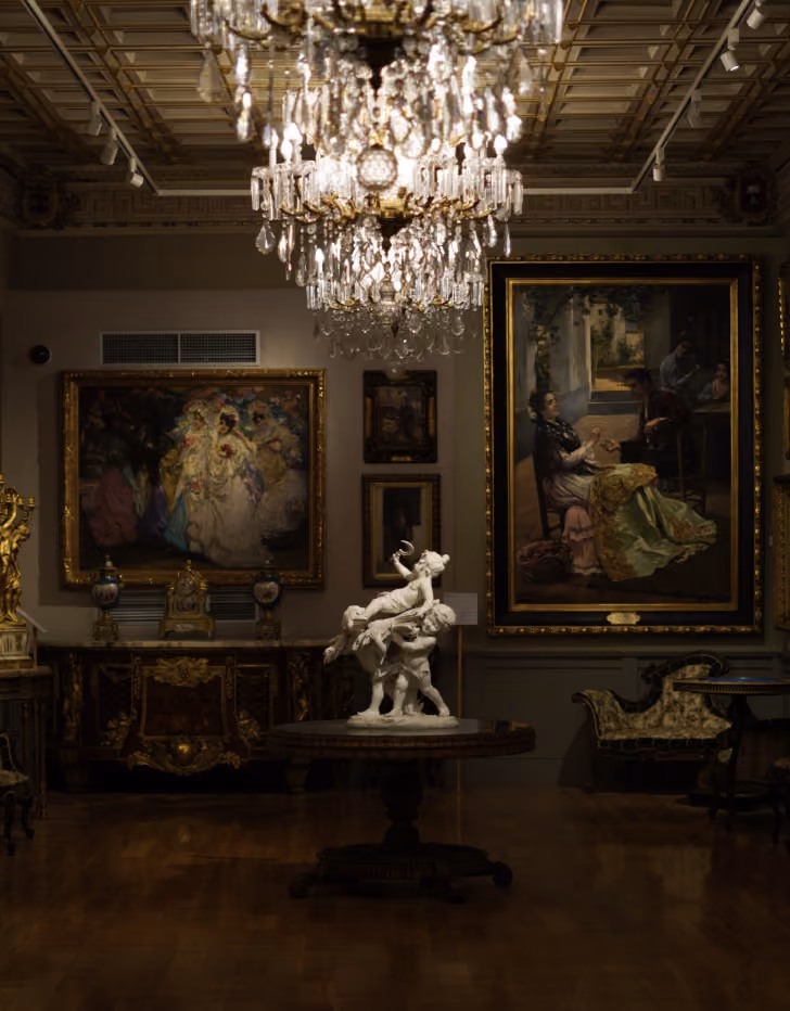 Intérieur d'une galerie d'art avec un lustre en cristal suspendu, une sculpture blanche au centre sur une table ronde, et des tableaux encadrés accrochés aux murs.
