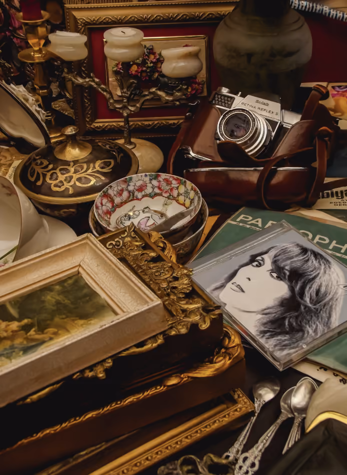 Collection d’objets vintage sur une surface, comprenant un appareil photo argentique dans un étui en cuir, plusieurs cadres dorés, des cuillères en métal, un bol décoré, un chandelier à trois bougies, ainsi qu’un magazine avec la couverture montrant un portrait en noir et blanc d’une femme aux cheveux longs.
