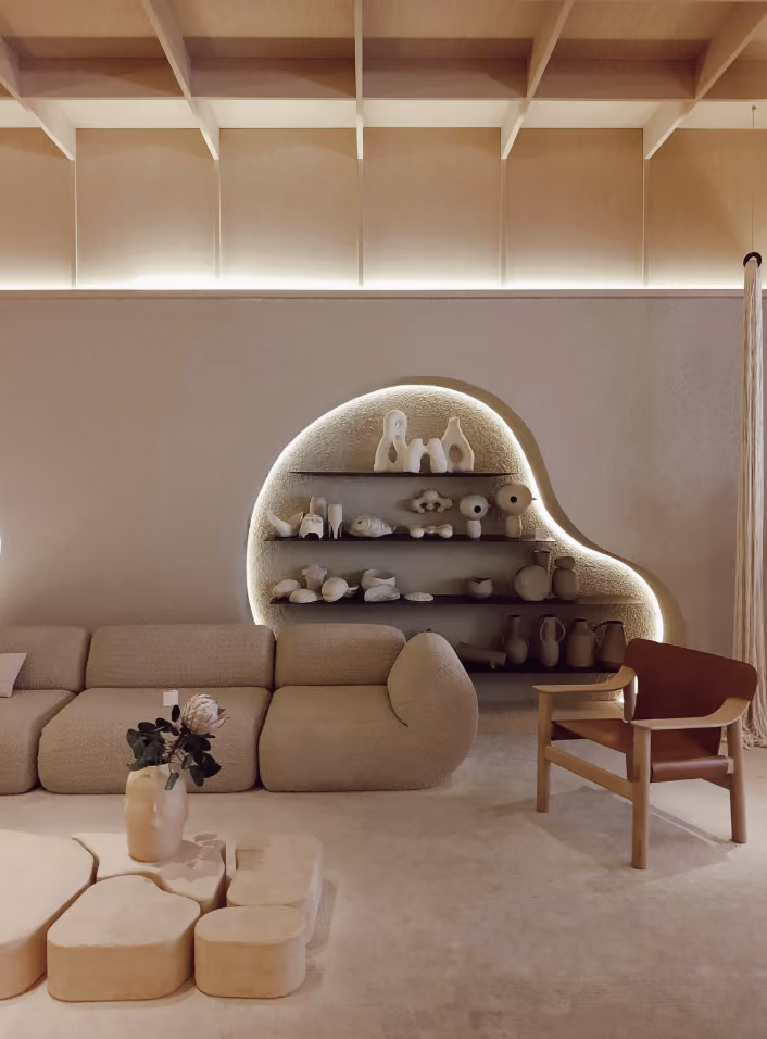 Salon moderne beige avec canapé aux formes arrondies, étagère intégrée encastrée avec éclairage LED et objets décoratifs en céramique, fauteuil en bois avec coussin marron et table basse organique avec vase et plante.