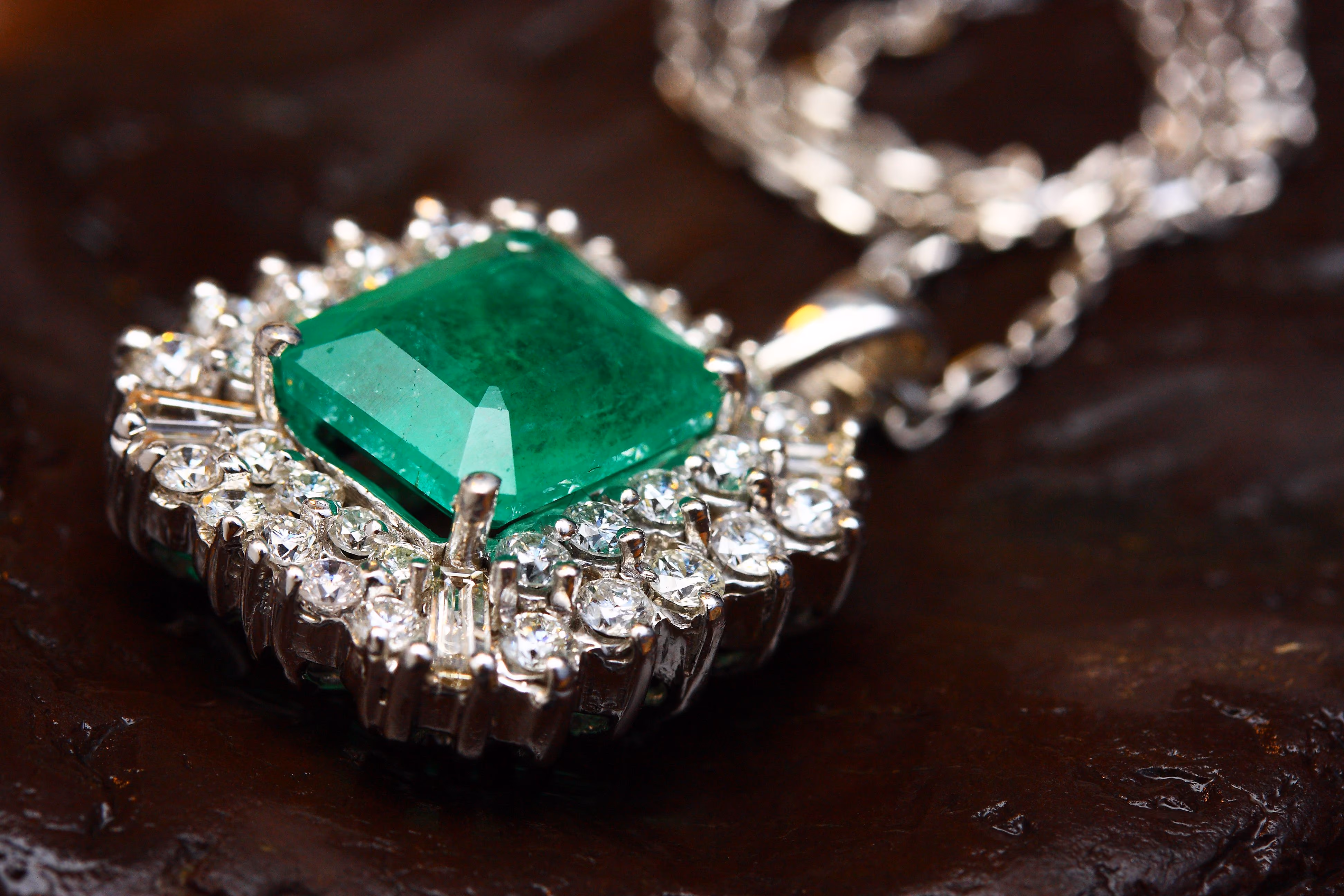 Pendentif avec une grosse émeraude carrée verte entourée de petits diamants brillants sur une chaîne en argent posée sur une surface sombre.