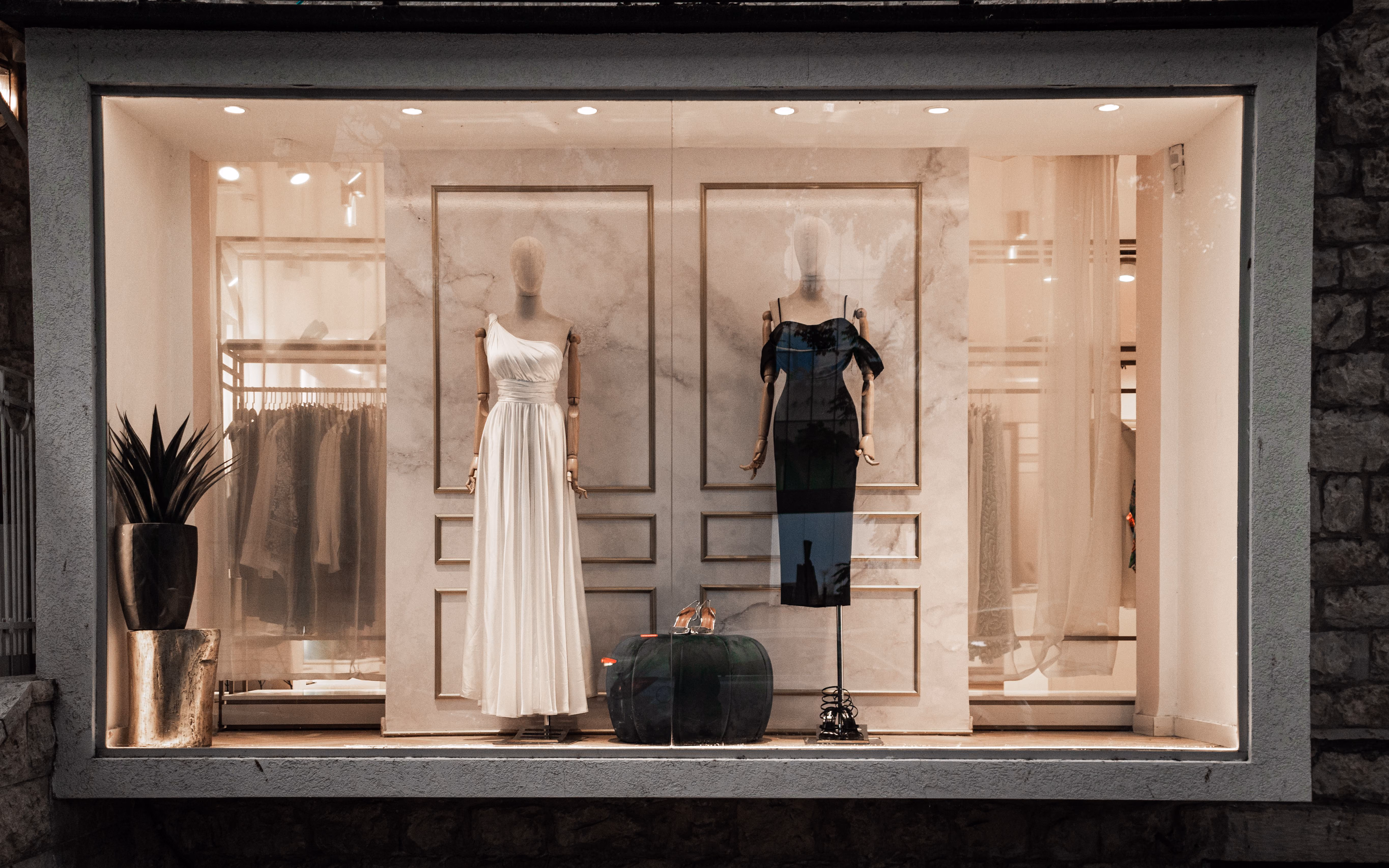 Vitrine élégante montrant deux mannequins, l'un portant une robe longue blanche sans manches et l'autre une robe noire épaule dénudée, avec une paire de chaussures à talons placée sur un tabouret entre eux, décor minimaliste avec un pot de plante à gauche.