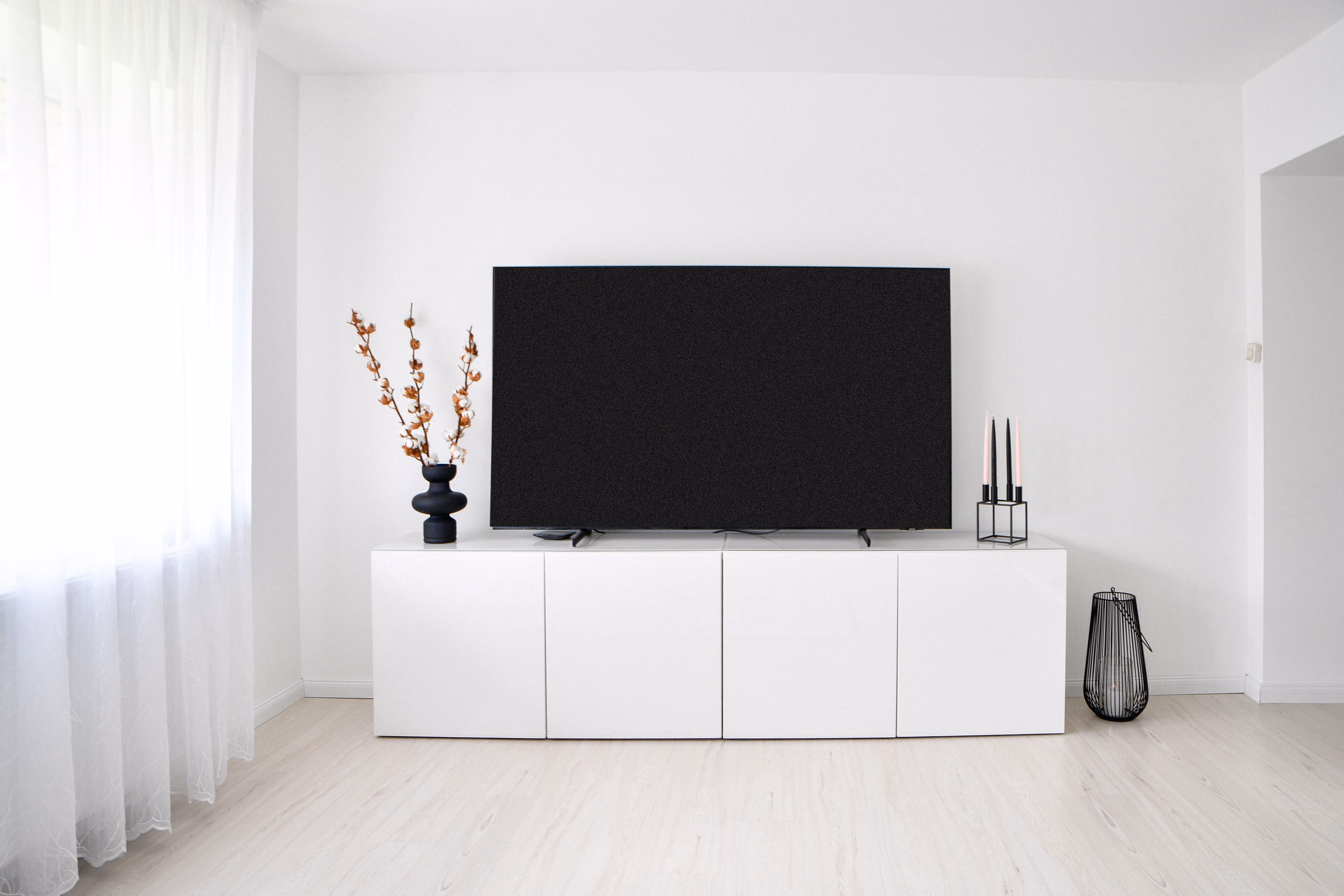 Salon minimaliste avec un meuble TV blanc, une grande télévision noire posée dessus, un vase noir avec des branches décoratives à gauche, deux bougies noires à droite, et une lanterne noire au sol sur la droite, éclairé par une fenêtre avec un rideau blanc à gauche.