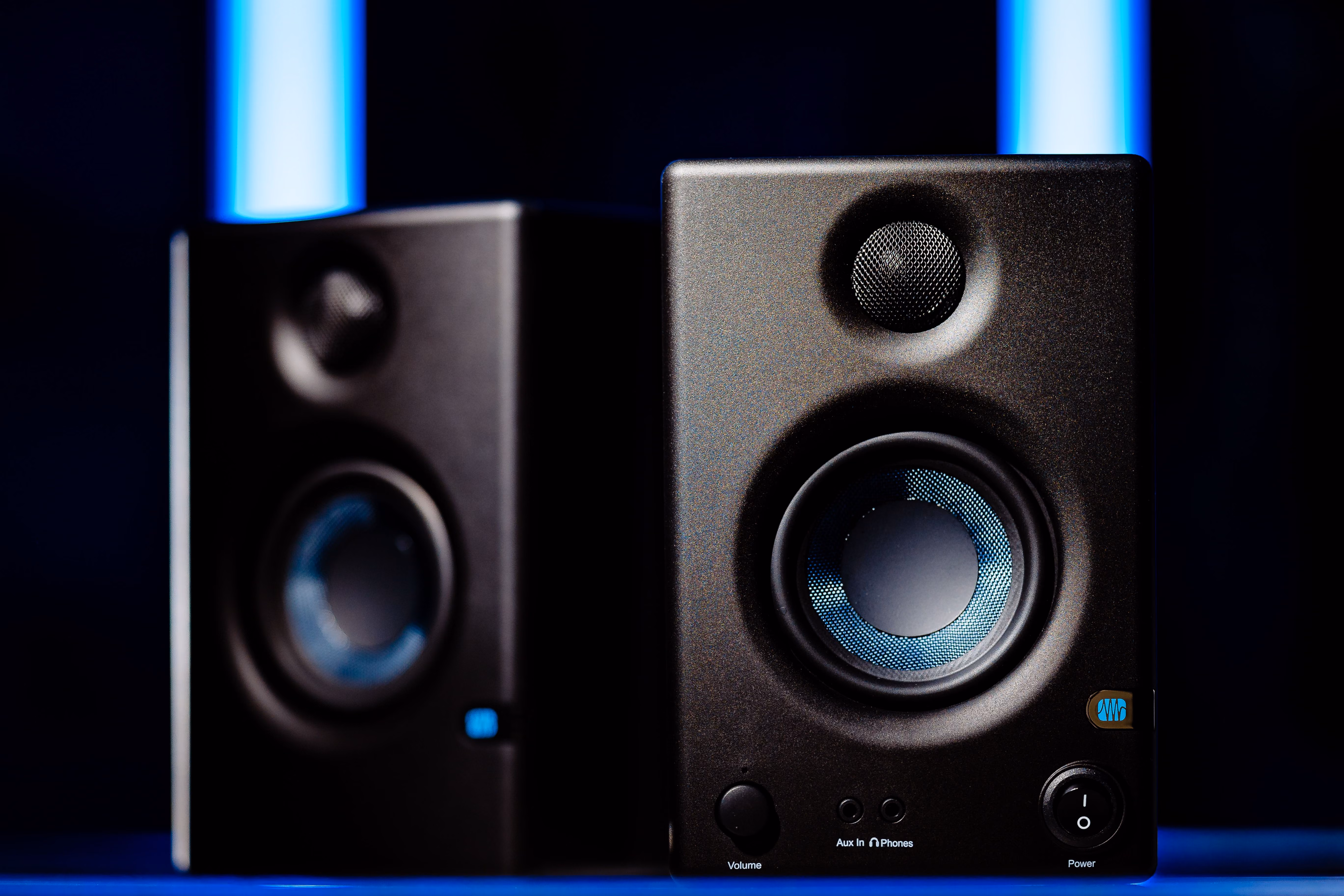 Deux enceintes de studio noires avec un cercle bleu lumineux sur chaque woofer, placées sur une surface avec un fond sombre et deux bandes lumineuses bleues verticales floues à l'arrière-plan.